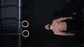 Vertical video. Caucasian male bodybuilder or exerciser is working out on a rings in the gym. The shadow of the athlete on the dark wall of the room.