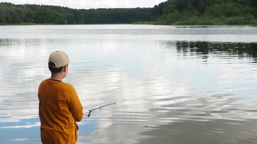 A little cute boy is fishing at a lake in summer. He holds the fishing rod bent in his hand and has a big catch. A fish jumps out of the water. The boy is surprised. active outdoor recreation