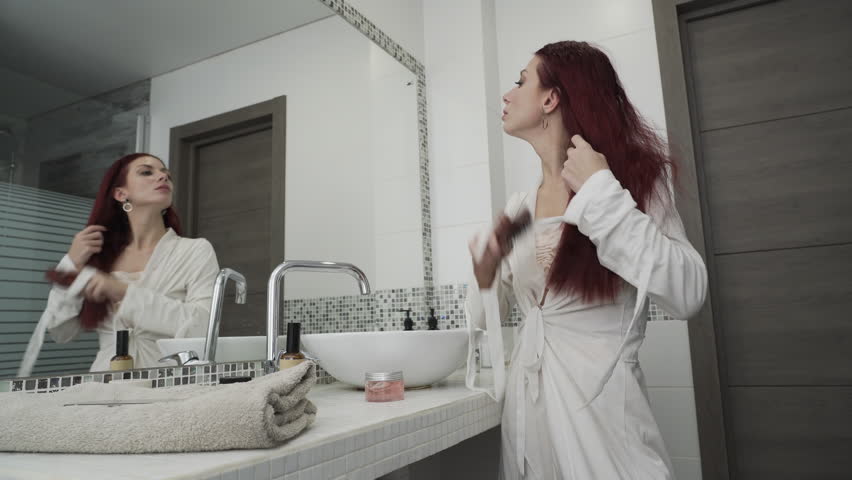 Woman combs her hair with a comb while standing near the bathroom mirror. Redhead woman takes care of her hair. Concept of a home hairstyle