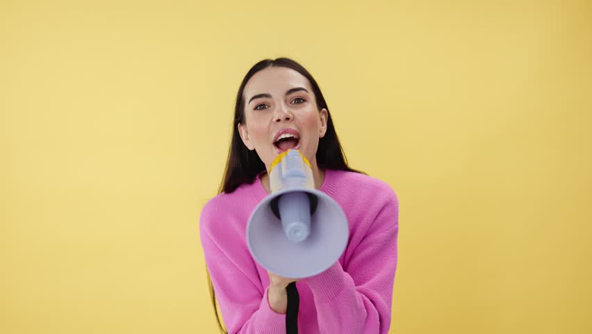 Cheerful female holding loudspeaker and telling announcement while making win gesture. Smiling woman talking in megaphone and supporting friends while standing in studio with yellow background. Royalty-Free Stock Footage #3437559733