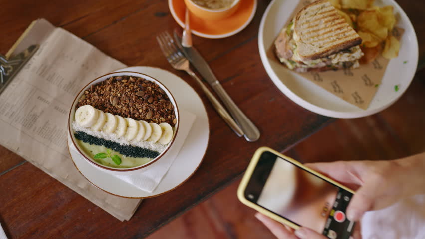 Female hands taking photos of smoothie bowl, menu on table by smartphone. Tasty sandwich, coffee on table. Woman photographs healthy food in hipster cafe on Bali. Blogger takes photo of the breakfast.