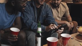 Medium shot of three multiracial male friends sitting on couch, drinking beer, man in middle showing video or photo to his friends, all laughing, Living room, stairs to first floor in backround