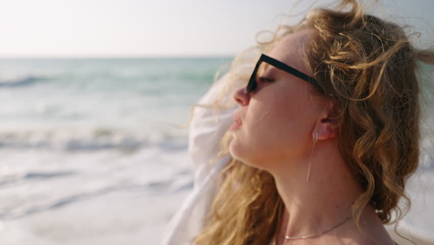 Pretty curly girl in sunglasses walking, touching hair at coast, sea surf. Young model posing, taking sun bath at ocean shore. Attractive woman enjoying summer vacation at the seaside. Close-up.