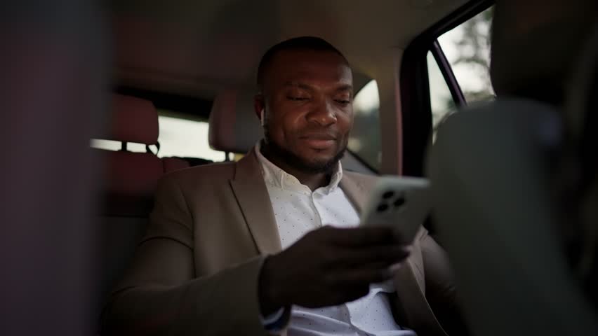 A confident man with Black skin in a brown suit with a short haircut and beard rides in the interior of a modern car and works on a white smartphone - Powered by Shutterstock - Get 15% off with code: PIKWIZARD15