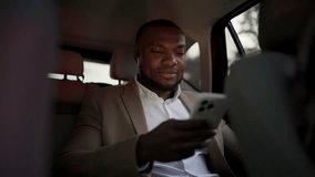 A confident man with Black skin in a brown suit with a short haircut and beard rides in the interior of a modern car and works on a white smartphone - Powered by Shutterstock - Get 15% off with code: PIKWIZARD15