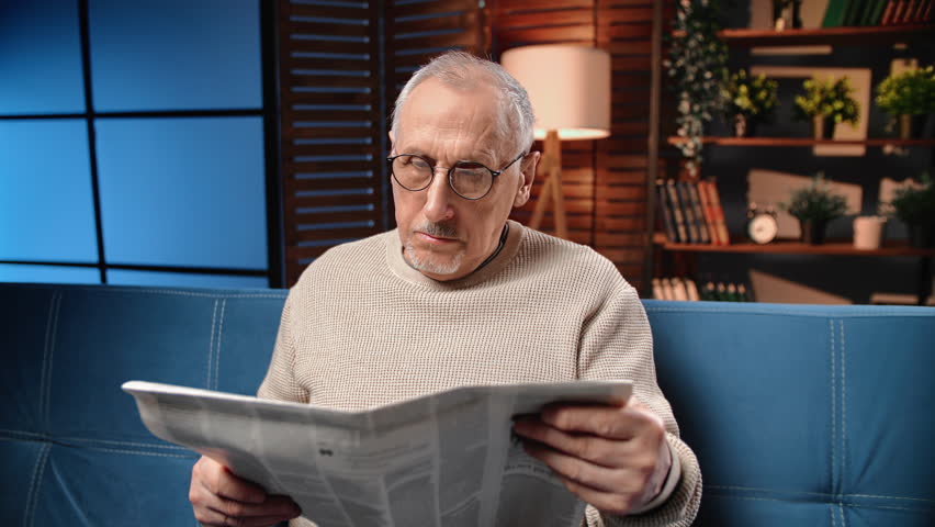 Worried old man reading newspaper and shaking head, portrait in living room of apartment in night. Bad news in world, financial and economic crisis, retired man examining daily newspaper, grandfather