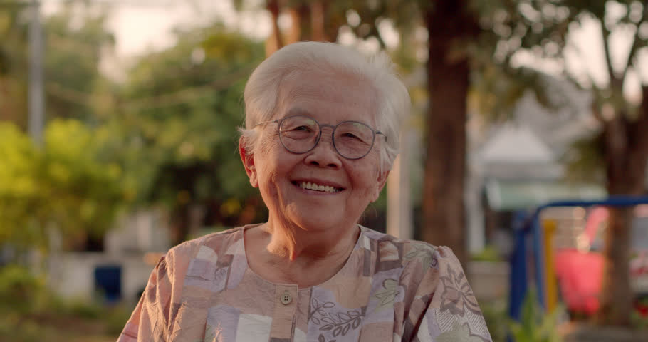 A smiling Asian woman who is an elderly person with white hair, wearing glasses, siting in a wheelchair in the park to rest and the sunset glowed golden in the evening.