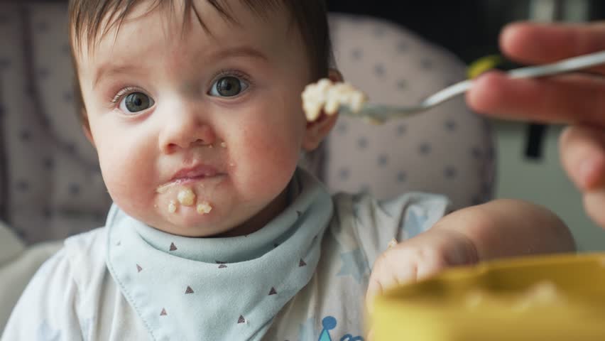 Happy family concept. close-up portrait of a baby sitting on chair and eating from a spoon. introduction of baby food in kindergarten.nanny feeds baby healthy food from box. breakfast in kindergarten Royalty-Free Stock Footage #3437940675