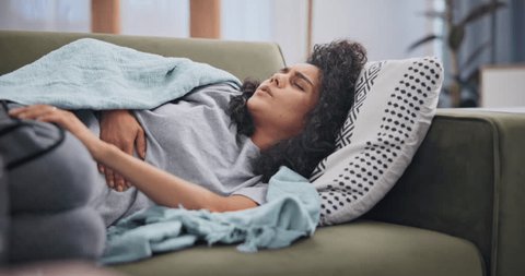 Home, stomach and period pain with woman on a couch, illness and sick in a living room. Person on a sofa, apartment and girl with a blanket and digestion issue with stress and constipation with gas 库存视频