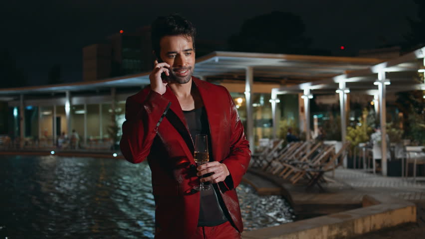 Glamorous guy calling smartphone at poolside party night close up. Smiling latin macho talking cellphone holding champagne glass outdoors. Happy businessman in fashionable red suit enjoy conversation. Royalty-Free Stock Footage #3438065633
