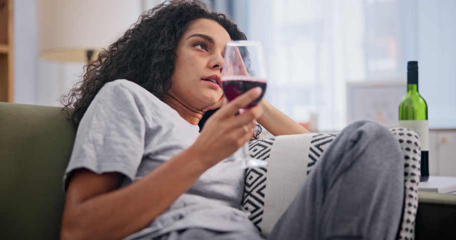 Depression, wine and drunk with bored woman on sofa in living room of home to relax or drink. Sad, frustrated or unhappy and young alcoholic person drinking in apartment with mental health problems