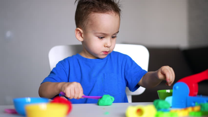 Calm focused baby boy in blue t-shirt playing at desk. Kid cuts the piece of green plasticine with a stick.