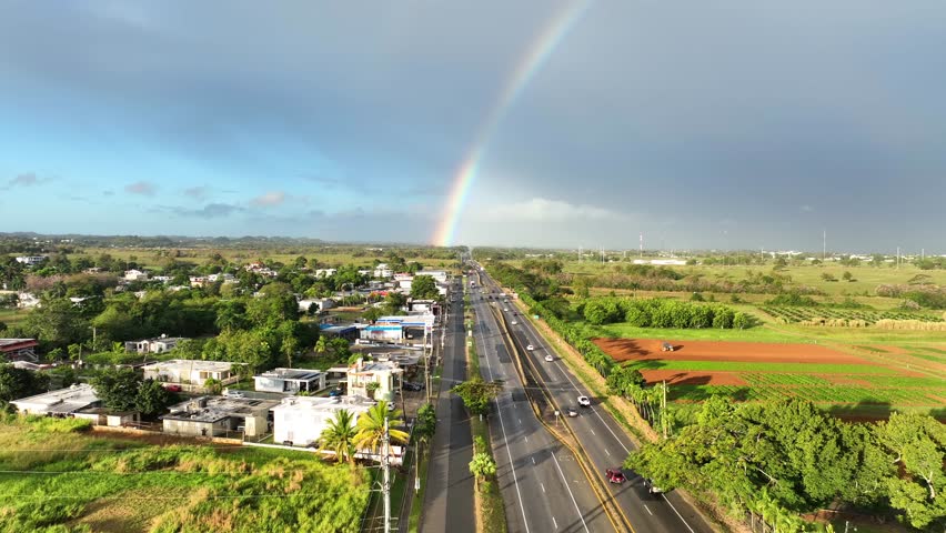 Highway in Aguadilla, Puerto Rico through rural countryside with cars driving towards rainbow in distance - Aerial 4K drone footage rising up