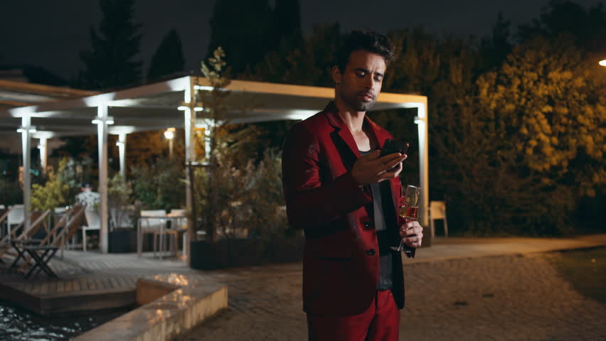 Successful gentleman talking cellphone at night weekend party. Confident spanish guy calling smartphone holding champagne glass outdoors. Smiling handsome businessman speaking phone in trendy red suit Royalty-Free Stock Footage #3438167705