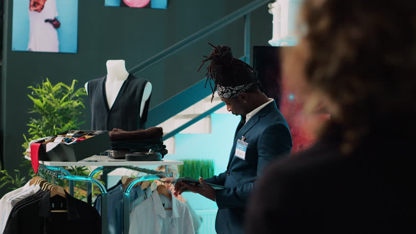 African american worker managing clothes stock on hangers, working on inventory using tablet in shopping center. Male store assistant in a suit preparing fashion collection on display. Camera A.