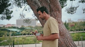 Relaxed tourist walking park holding beverage takeaway close up. Side view attractive hispanic guy strolling green area looking smartphone screen. Calm carefree man enjoy weekend rest with coffee cup. - Powered by Shutterstock - Get 15% off with code: PIKWIZARD15