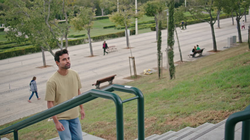 Relaxed guy going upstairs city park at carefree summer weekend. Positive italian man walking up stairs urban street alone looking around. Stylish handsome tourist exploring calm green town district. Royalty-Free Stock Footage #3438178085