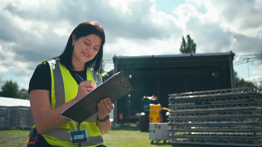Female event production manager with radio headset wearing hi-vis safety vest and using clipboard setting up stage for outdoor music festival or concert - shot in slow motion