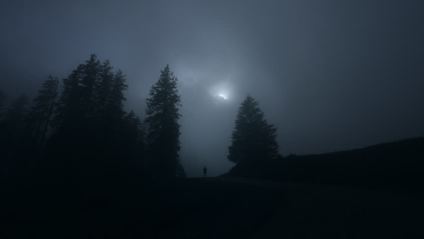 Silhouette of a hiker walking in misty hills surrounded by pine trees at night with moon in the sky