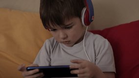 Child Happy Playing Games With Phone. Little Boy With Headphones Playing Video Game on Mobile Phone. Preschooler Plays Video Game On Smartphone. Harm for Mental Kid Health Eyesight Gambling Addiction