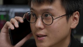 Young Asian businessman in glasses engaged in a phone conversation with blurred stock market screens in background, close-up - Powered by Shutterstock - Get 15% off with code: PIKWIZARD15