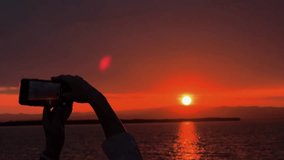 Close-up of woman's hands holding smartphone and taking pictures or video of amazing orange sunset at the sea. Beautiful panoramic view of sun over the mountains.