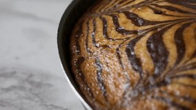 Zebra cake with chocolate stripes pattern baked in a metal cake pan, placed on a white marble tabletop, close-up panning shot video.