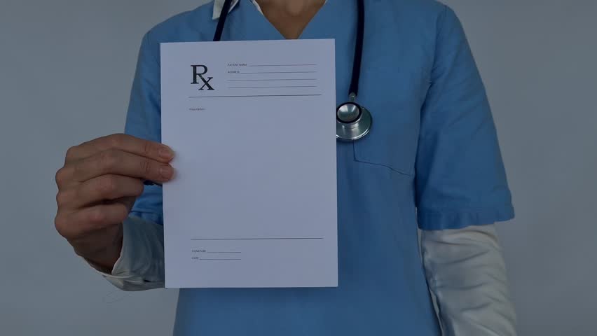 A female doctor in a blue uniform is holding a blank prescription pad and pen. She is looking at the camera with a serious expression Royalty-Free Stock Footage #3438739739