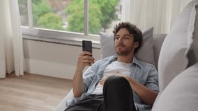 Satisfiedcaucasian guy is enjoying watching videos on couch. Carefree calm white man watch videos on phone, resting on sofa at home 
