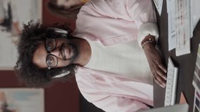 Vertical Waist up slowmo portrait of Biracial male interior designer in wireless headphones sitting at desk with blueprints on it and smiling at camera, working from home office - Powered by Shutterstock - Get 15% off with code: PIKWIZARD15