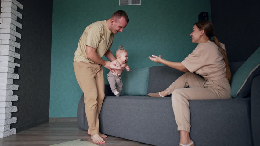 Father is holding a little baby in grey pants who steps by the sofa. Infant walks to his mom sitting in front.