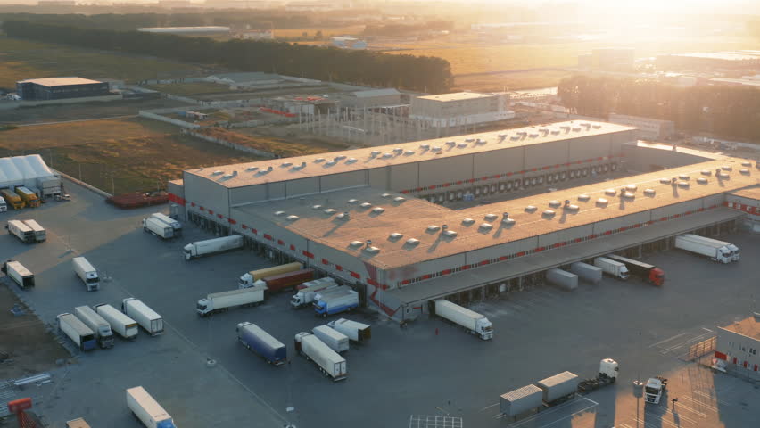 Aerial all-round view of a logistics park with a warehouses. Loading hub with many semi trucks with cargo trailers standing at ramps for load and unload goods at sunset