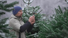 Male weblogger shooting video using camera phone for social media networks and followers on online streaming services about selecting fir trees at a Christmas market in winter snowy days. 