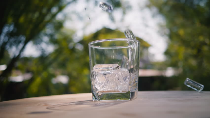 slow motion view of pouring ice tube into glass of water on table with a natural background, low angle close up with an orbit rotation camera view Royalty-Free Stock Footage #3438974987