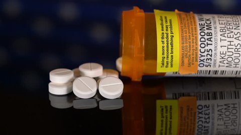 Sideways pan on oxycodone, the generic name for a range of opoid pain killing tablets. Prescription bottle for Oxycodone tablets and pills on wooden table with USA flag in background