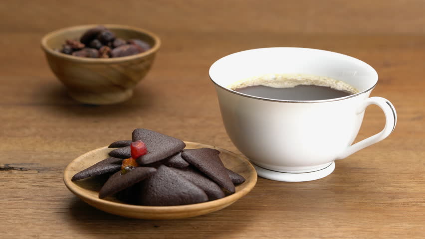 Lifting white ceramic cup of hot black coffee, drink and placing it on a wooden board with delicious homemade dry mixed fruit brownie crackers in wooden board and cacao nibs in a cup.