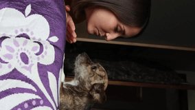 Caucasian girl playing with her puppy on the bed inside the house, while the dog kisses her on the face, vertical