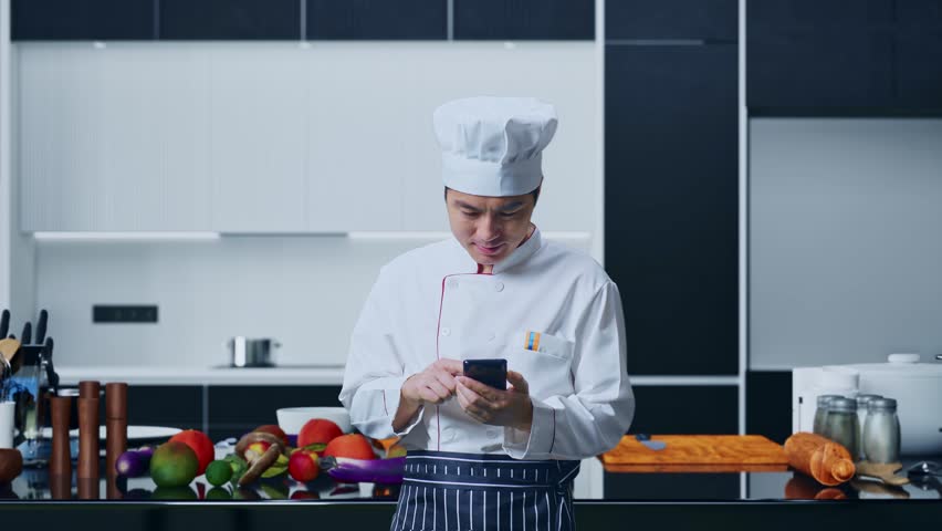 Asian Man Chef Smiling And Pointing To Smartphone In His Hand While Standing In Home Kitchen - Powered by Shutterstock - Get 15% off with code: PIKWIZARD15