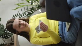 Vertical video of focused young woman using computer, typing or searching prescription on medicine label about vitamins information online sitting on couch at home. Wellness and dieting concept
