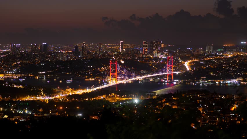 Night timelapse over Istanbul, capturing vibrant city lights and iconic Bosphorus Bridge. Road glows like winding river, passing through red pillars of bridge. Illuminated boats cruise on dark water