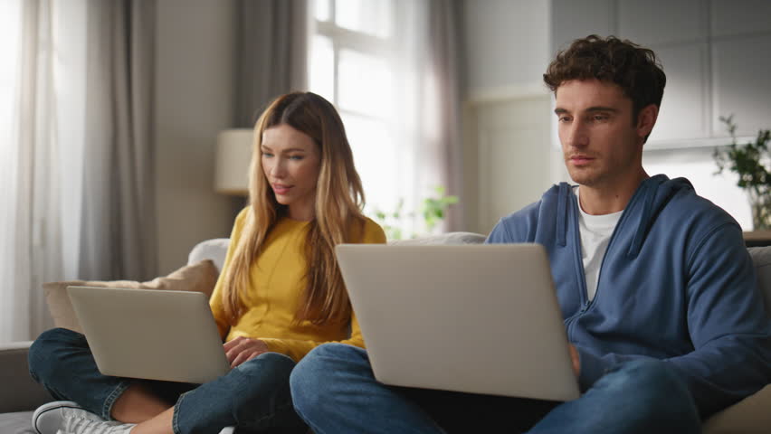 Cheerful sweethearts browsing laptops at couch closeup. Happy man positive woman typing keyboard looking each other with smile. Young couple working computers together at light apartment interior Royalty-Free Stock Footage #3439607525