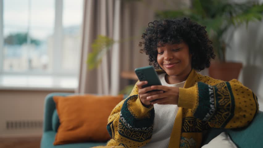 Young woman sitting with phone. African American woman hold in hand use mobile phone typing messages. Girl spend free spare time in living room. Communication on social networks, rest concept. - Powered by Shutterstock - Get 15% off with code: PIKWIZARD15