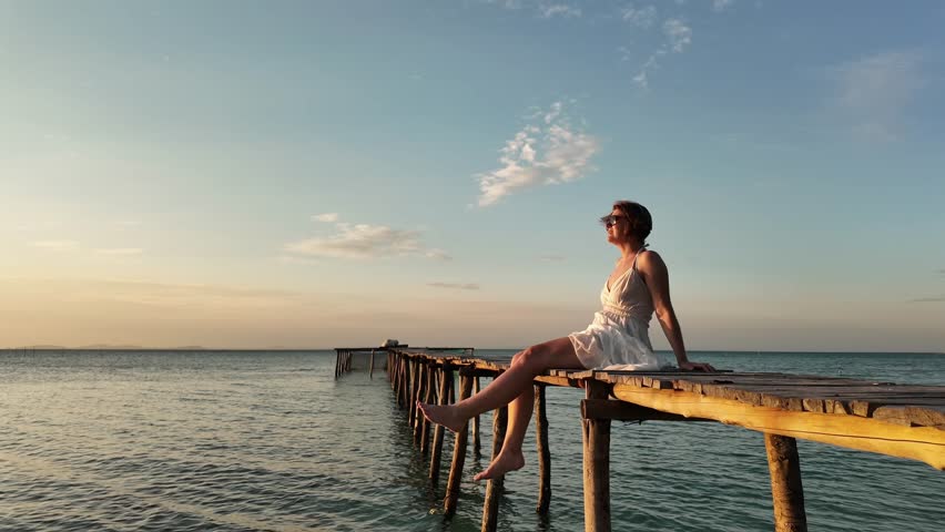 Serene woman in a white dress sitting on a wooden pier over the sea at sunset, symbolizing peacefulness and summer vacations