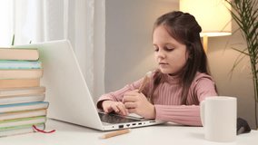 Learning remotely using a laptop. Table setup for online learning. Kid attending a virtual lesson. Caucasian little brown haired girl using laptop writing down task studying online at home