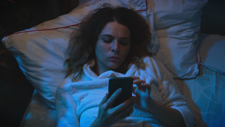 Insomnia concept. Sleepless tired young woman lying in bed scrolling smartphone at night. Unhealthy lifestyle.