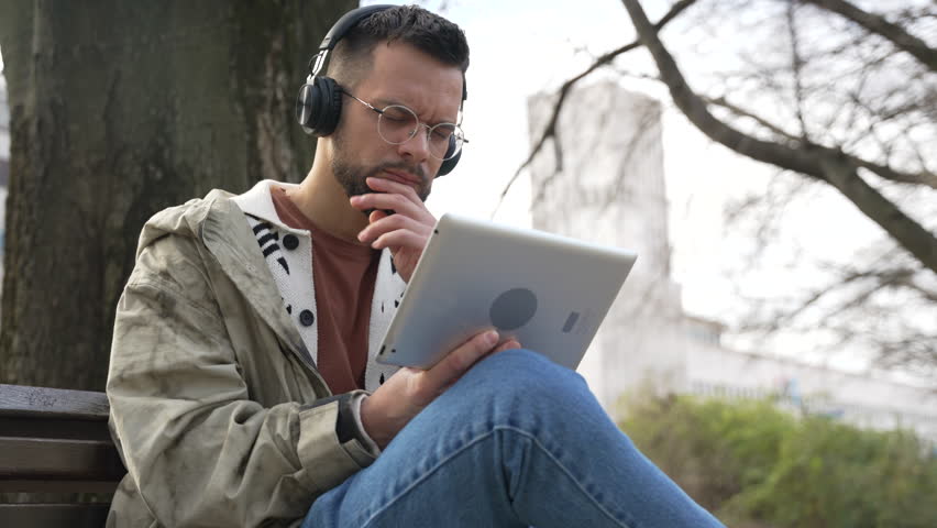 Young freelance expatriate hipster generation Z man sitting in park relaxing reading emails checking business economy statistics getting nervous. Serious businessperson small company owner on vacation