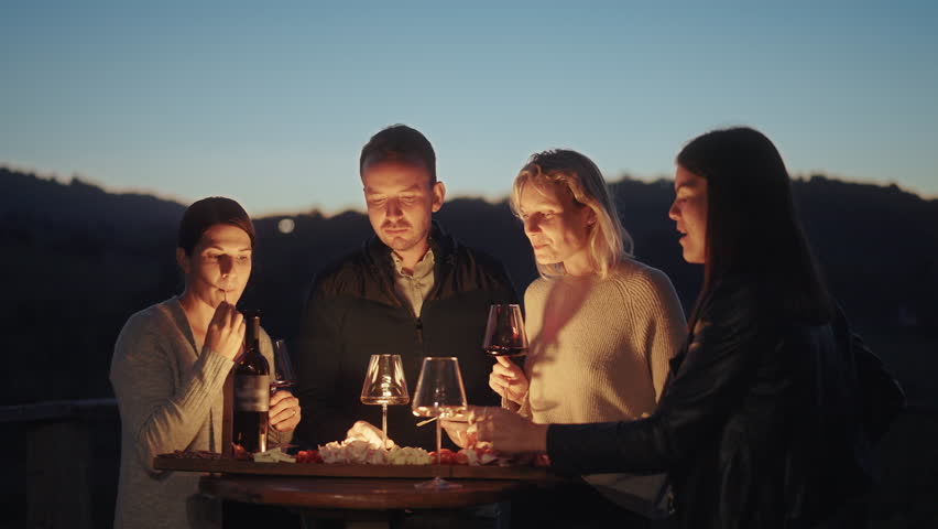 Group of friends enjoying wine tasting with local cheese, prosciutto and wine at sunset. Countryside vineyards and mountains in the background. Happy adult friends having fun, drinking wine and eating