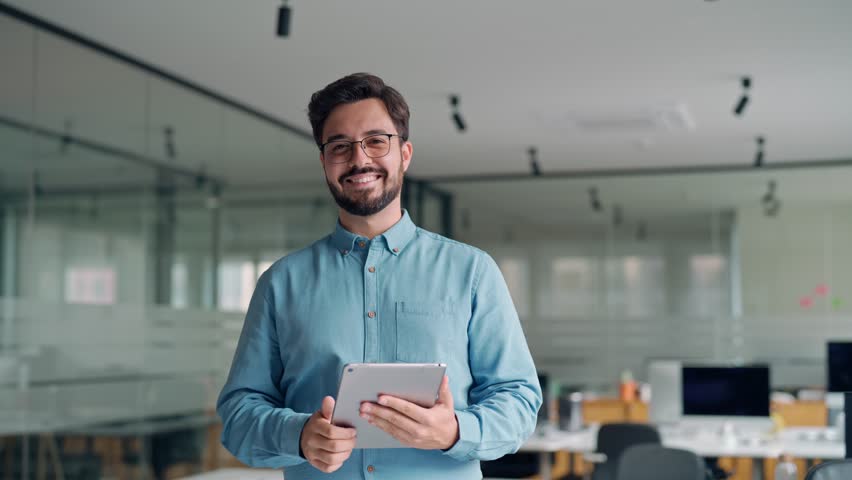 Smiling busy young latin business man manager using tablet computer, happy hispanic businessman executive looking at camera holding tab working standing in office. Portrait. - Powered by Shutterstock - Get 15% off with code: PIKWIZARD15