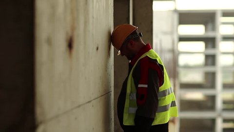 Workman knocks his head against the wall. Stressed worker in hard hat stands on construction site and knocks his head against the concrete wall, self-condemnation and self-torture