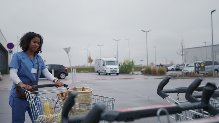 Female doctor grocery shopping after work. Beautiful healthcare worker pushing shopping cart with groceries across store parking lot. Work-life balance of healthcare worker as parent and partner.
